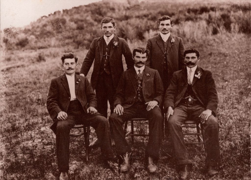 Five men in a sepia photograph pose for their photo sat on chairs in a field in 1910