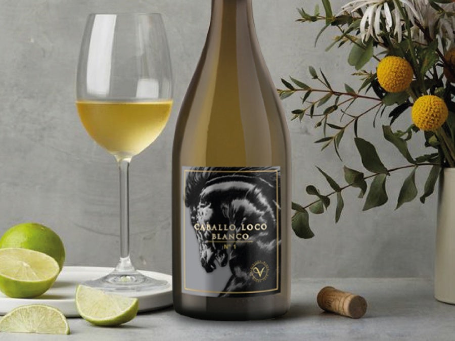 A close up of a wine label reading Caballo Loco Blanco sat on a table next to a glass of white wine, slice of lime, wine cork and miniature orange tree
