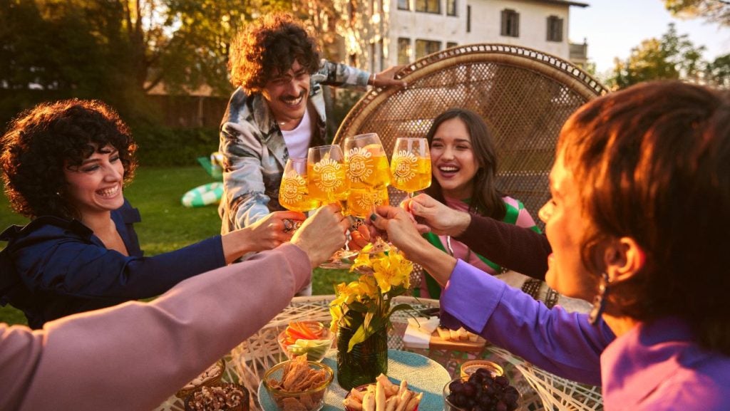 group of friends outdoors clinking glasses of orange-coloured beverage, house and leafy garden in background.