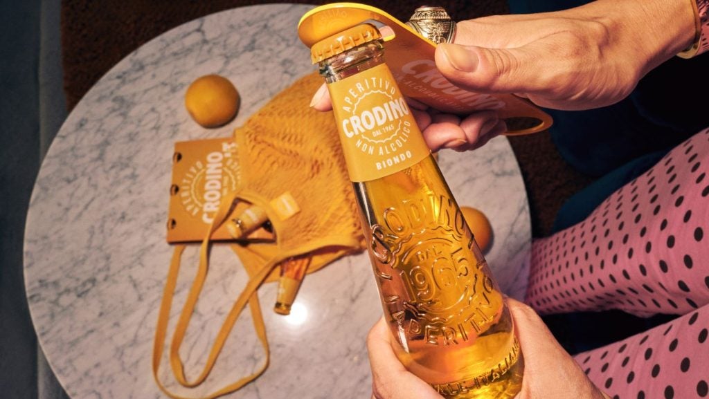 person in pink polka dress opens bottle of Crodino-branded orange drink in bottle, over marble table with orange and orange Crodino-branded bag.