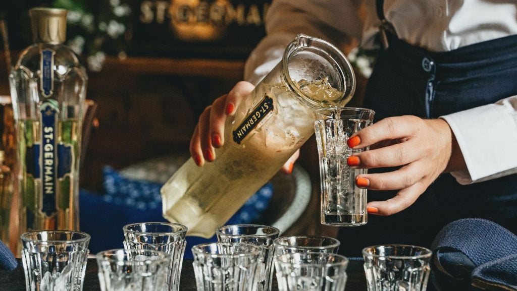 Woman with orange painted nails pours drink with ice out of St Germain branded jug into glass. Selection of glasses in foreground. St Germain liqueur in gold and glass branded bottle in background,