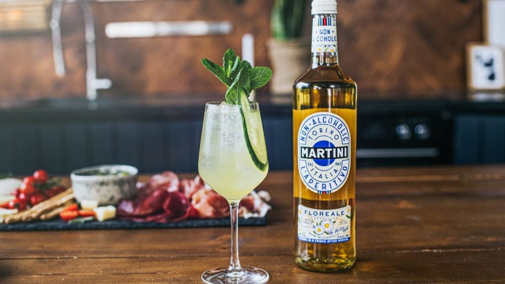 Martini Floreale non-alcoholic spirit, yellow hue spirit in bottle on wooden table, next to cocktail in glass with cucumber and mint sprigs. Charcuterie stone board in background.