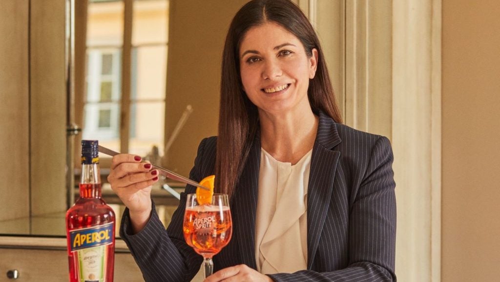 women in black blazer, black hair and white shirt holding orange drink with Aperol bottle, indoors.