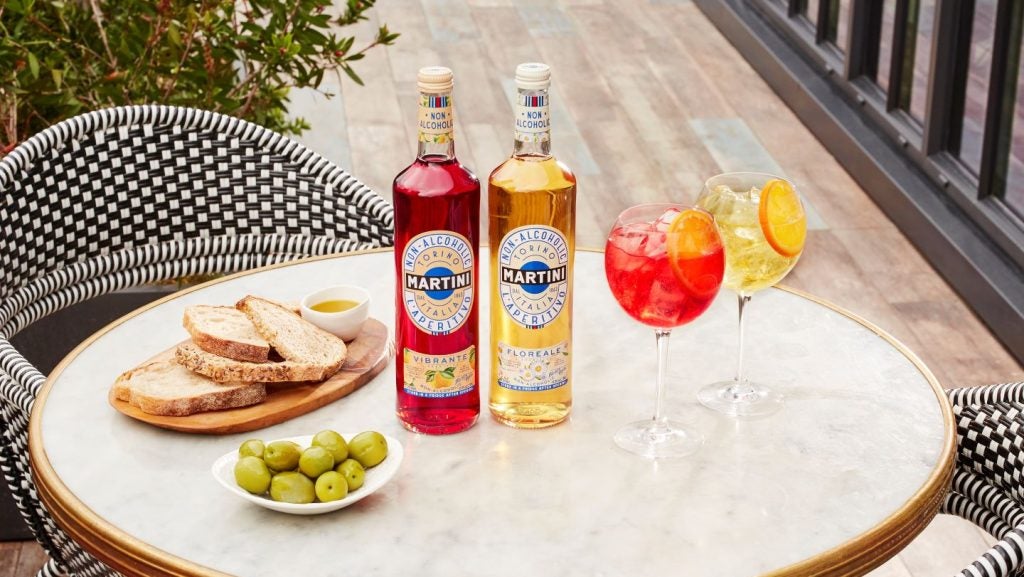 Two Martini branded non-alcoholic spirits, yellow and red colour, alongside two full glasses, plate of bread and olive oil, and plate of olives on marble table with black and white chairs, outdoor setting.