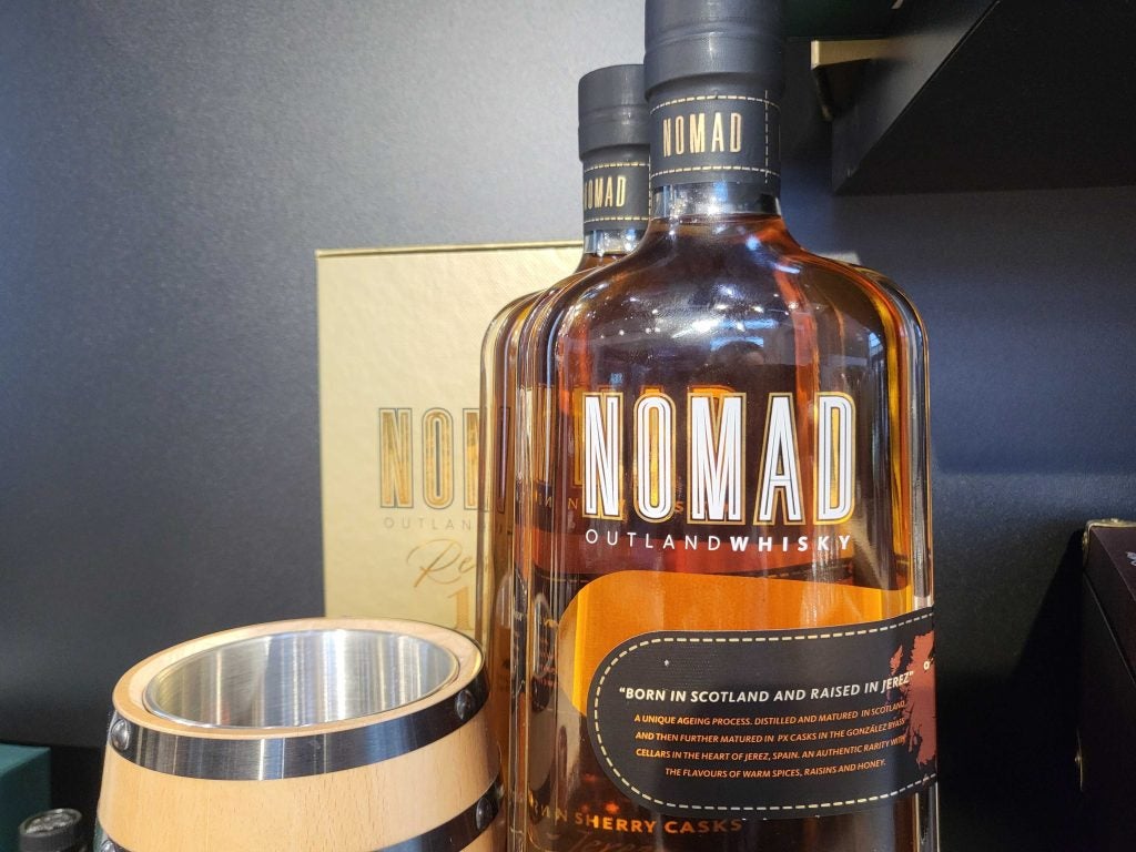 A bottle of Nomad whisky next to a wooden cup on a black shelf