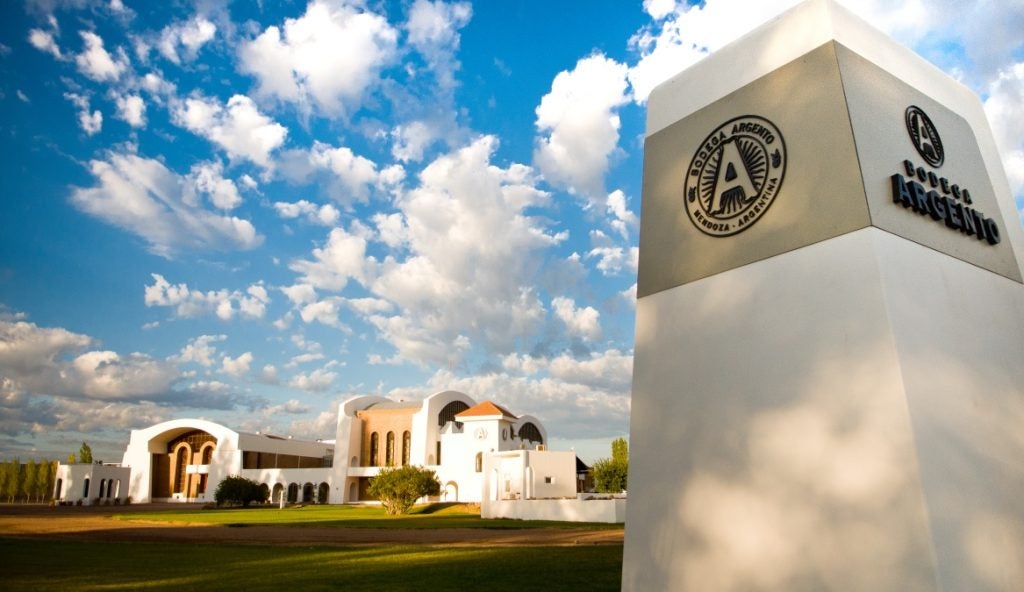 A white pillar with a sign saying Bodega Argento in the forefront, with a winery, green grass and blue cloudy sky in the backgrond