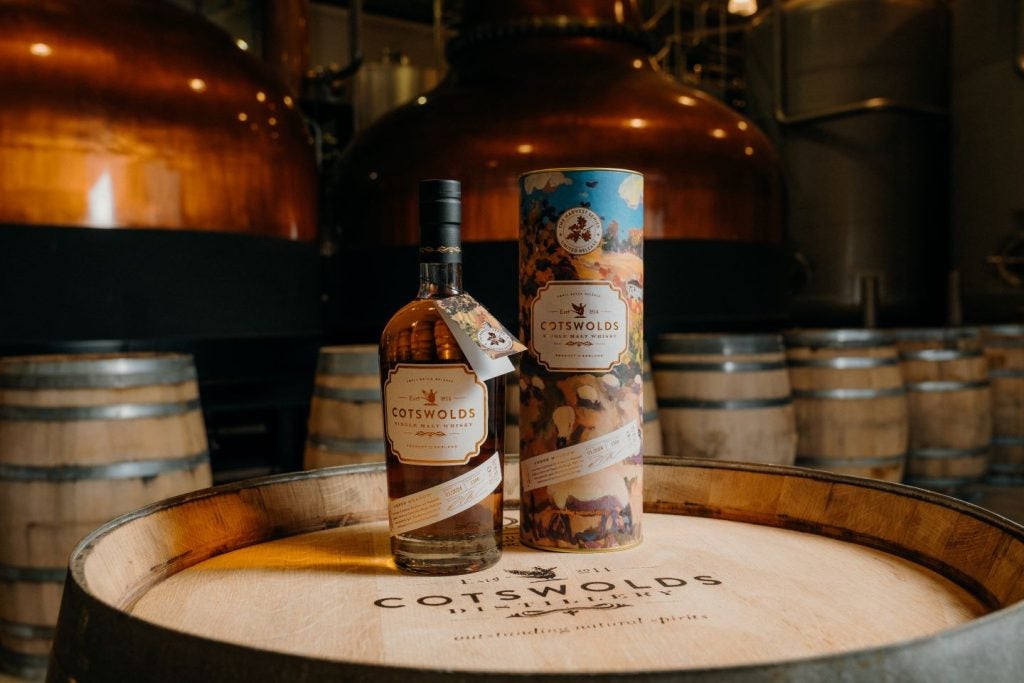 Bottle of whisky and packaging on barrel with Cotswolds Distillery brand, indoors.