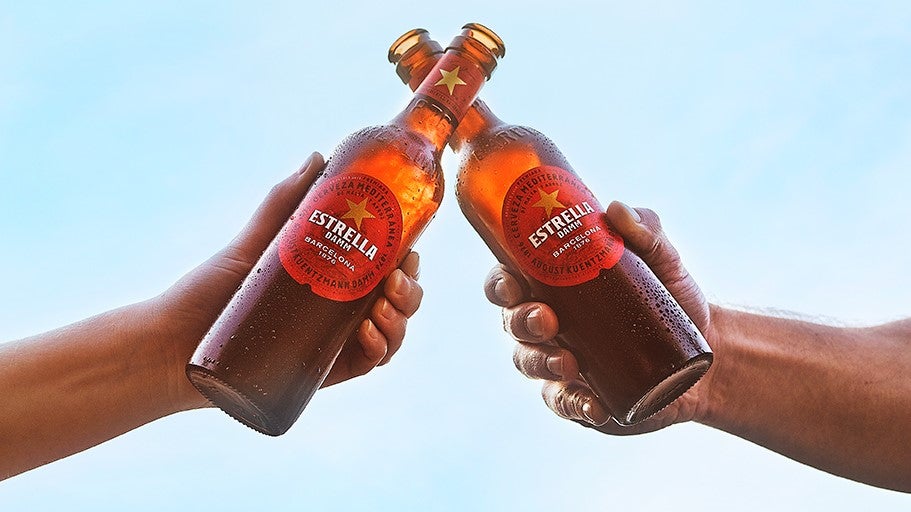 two people clinking Estrella Damm bottles together against blue sky