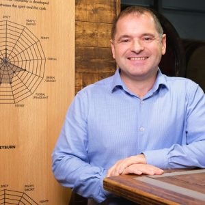 Man in blue shirt smiling, standing next to wooden table.