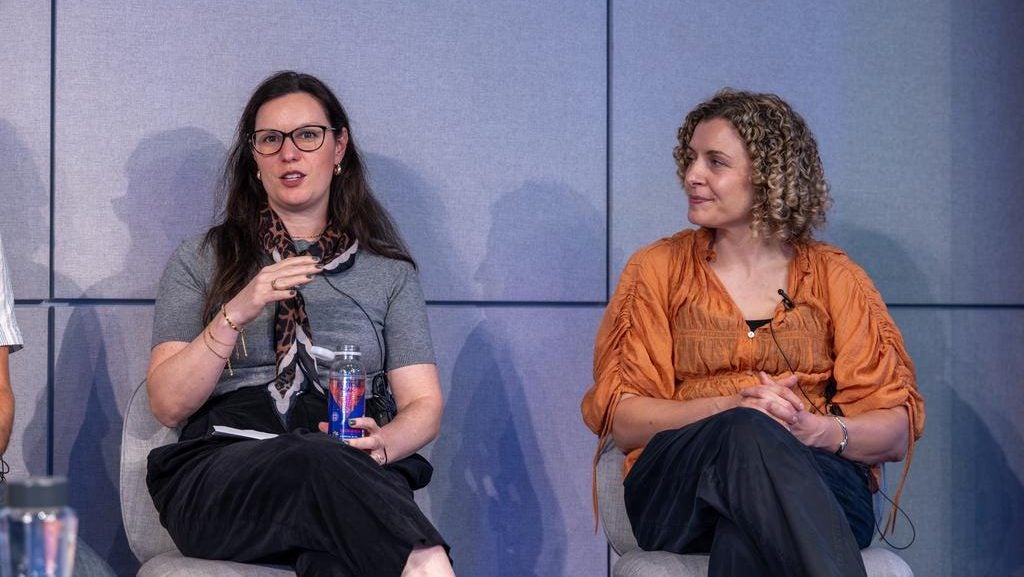 Two women sitting on stage, one speaking with microphone.